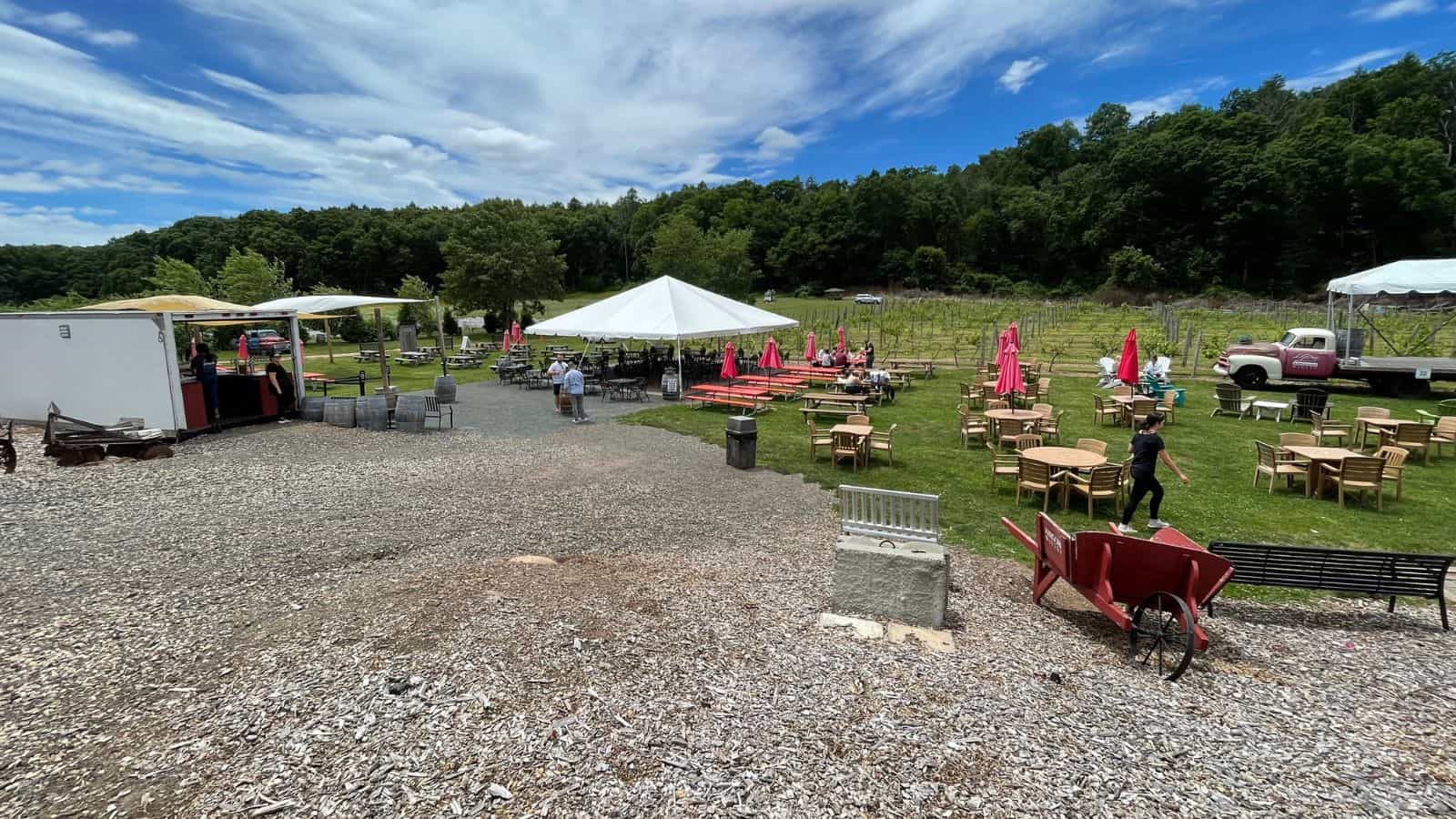 Outdoor seating area with round tables, red umbrellas, a white tent, people walking, a food truck, and a vintage truck, surrounded by trees under a partly cloudy sky.