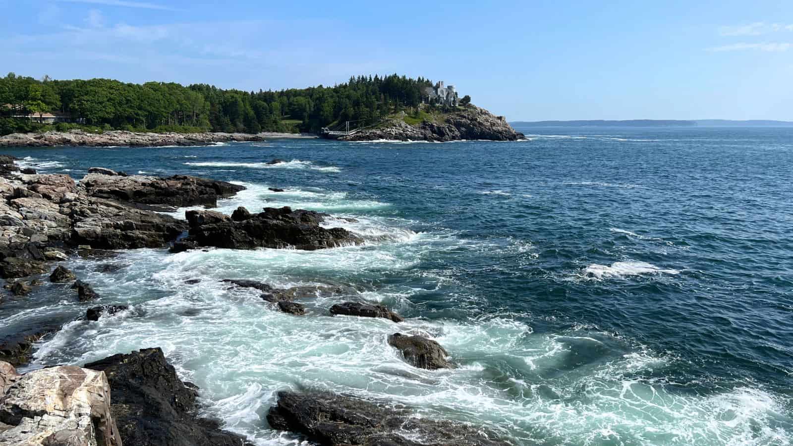 Rocky coastline with waves crashing on the shore, a forested hill in the background, and a clear blue sky above.