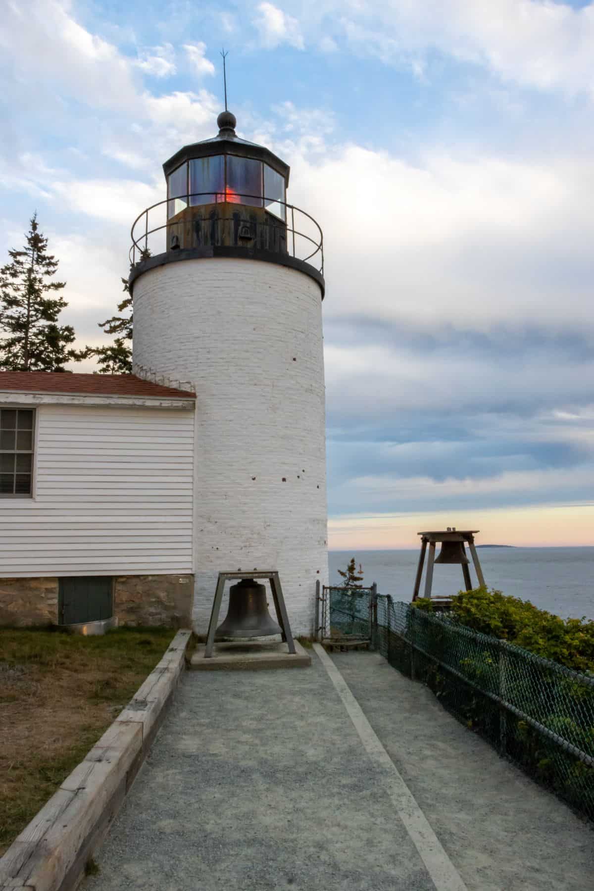 A white lighthouse with a red light sits near the edge of a cliff overlooking the ocean, with trees and a cloudy sky in the background.