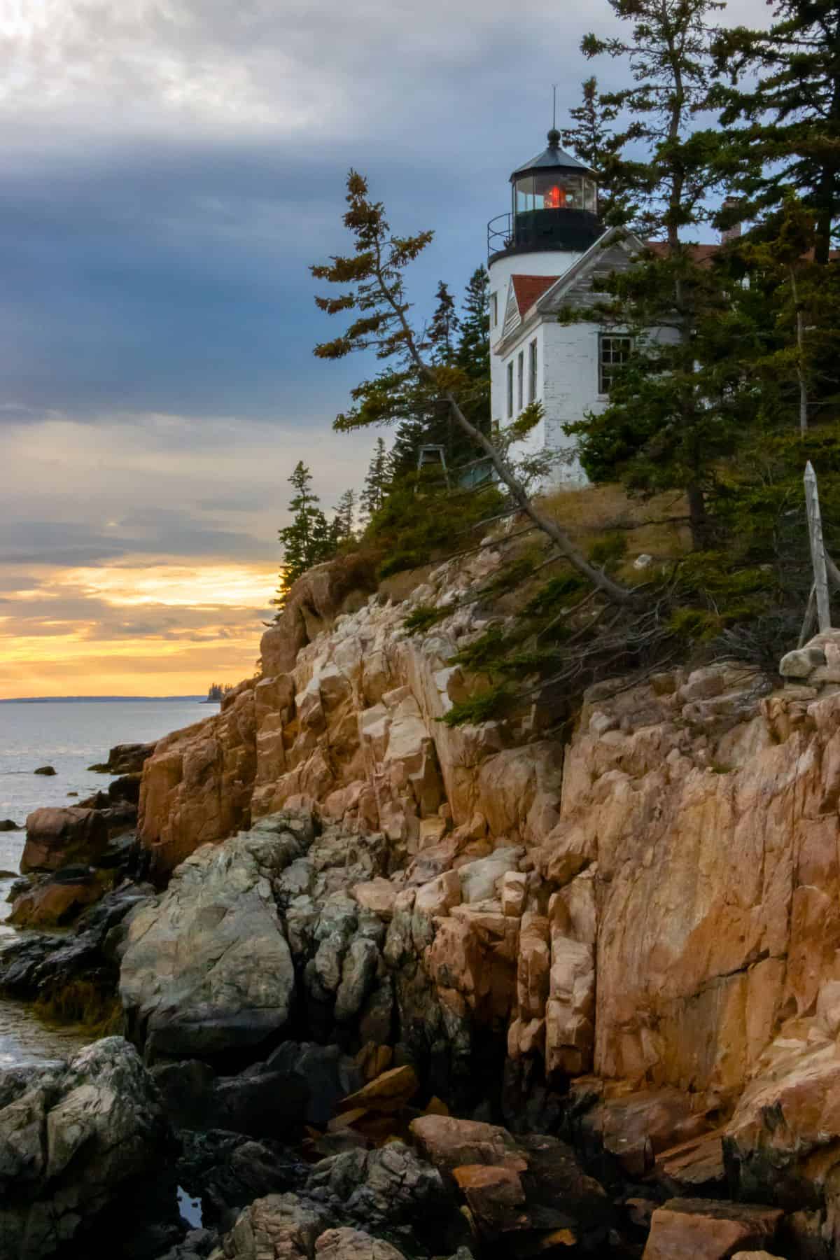 A white lighthouse sits atop rocky cliffs surrounded by pine trees at sunset, overlooking the ocean under a partly cloudy sky.