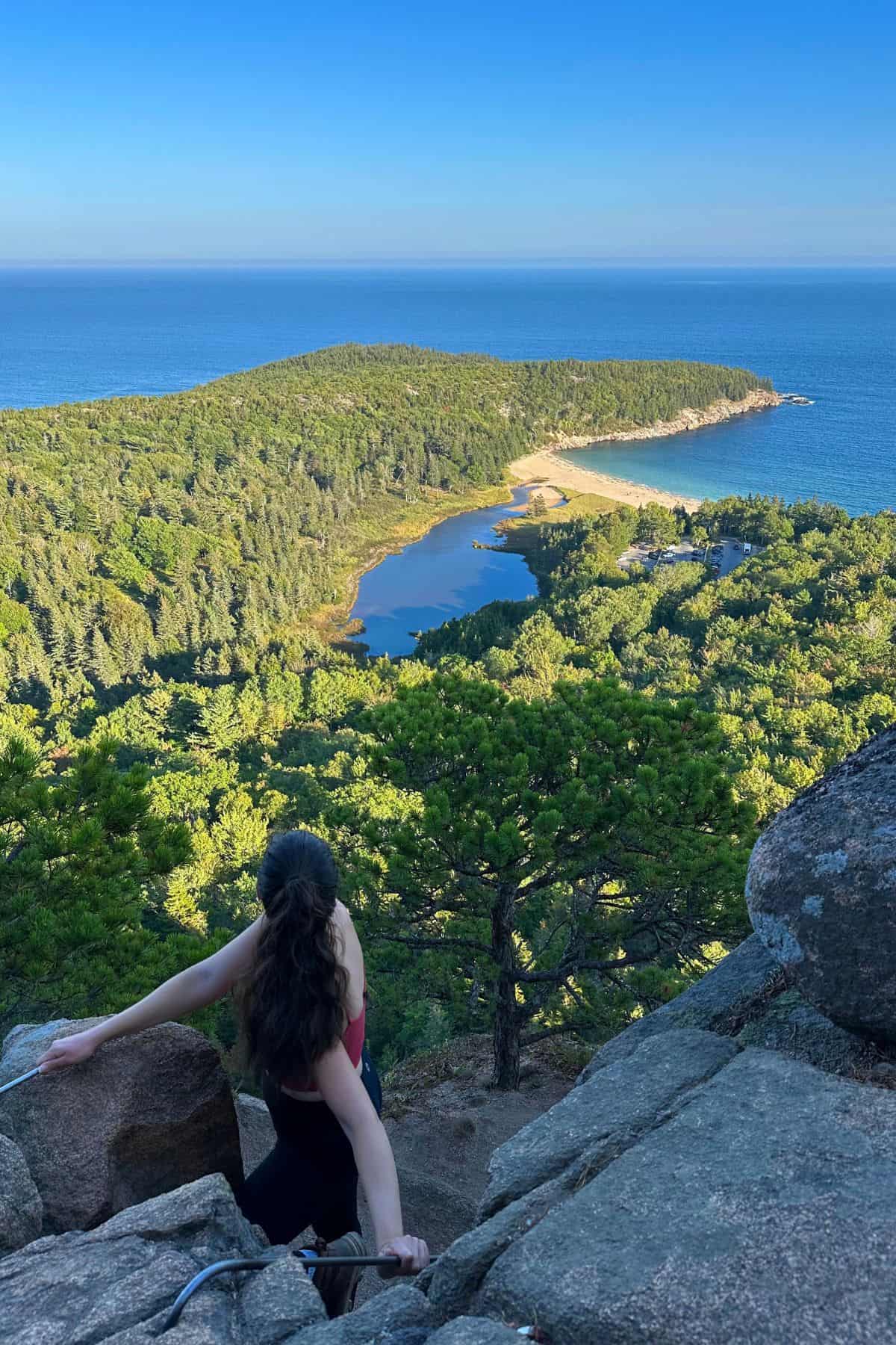 A person with long dark hair climbs rocks overlooking a forested peninsula, a beach, and the ocean under a clear blue sky.