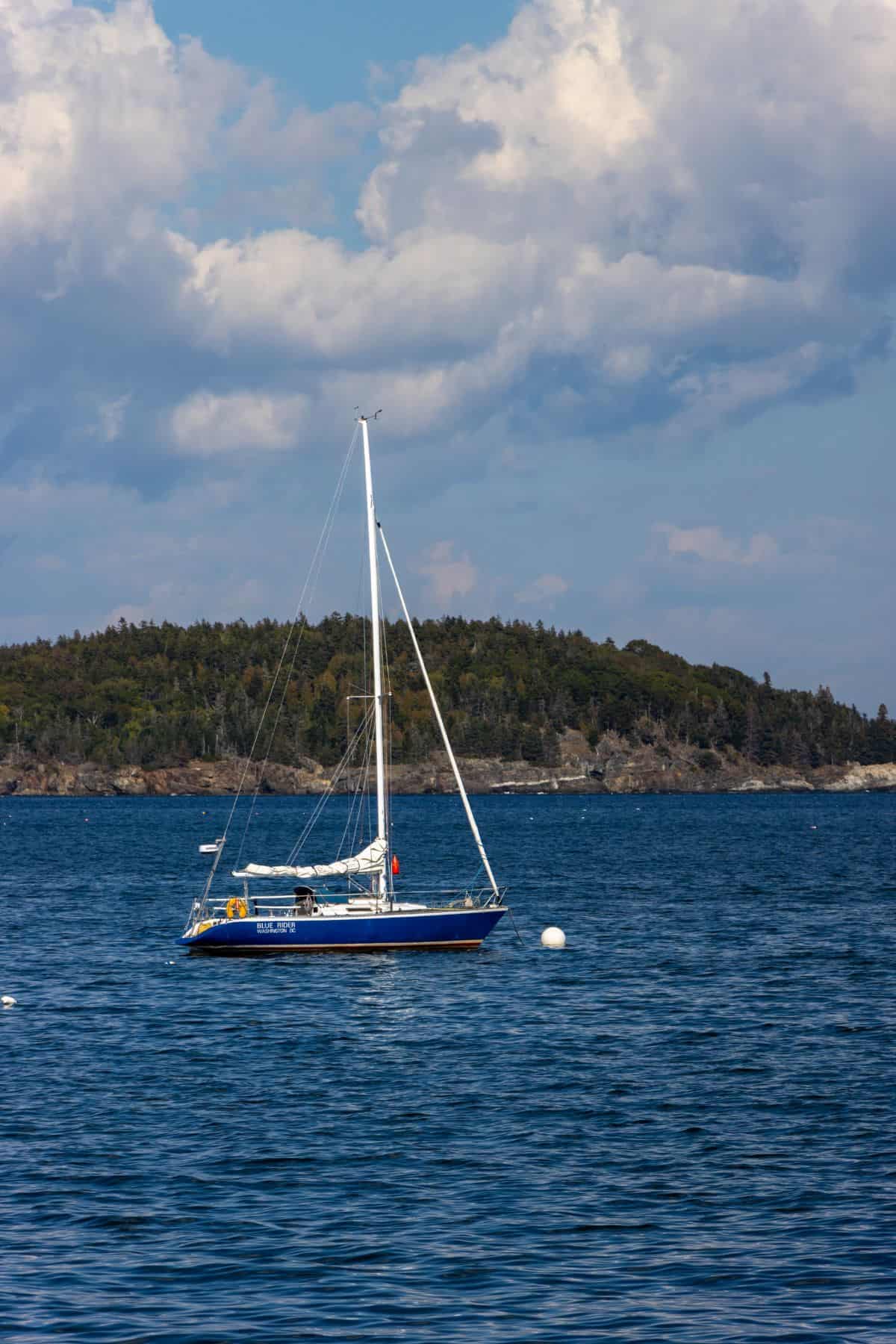 A blue sailboat is anchored on calm water near white buoys, with a forested shoreline and a partly cloudy sky in the background.
