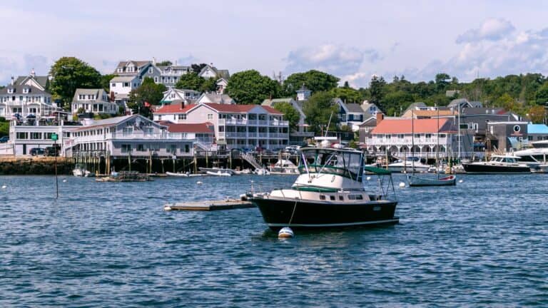 A small motorboat floats on calm water near a dock, with coastal buildings and trees lining the shore in the background under a partly cloudy sky.