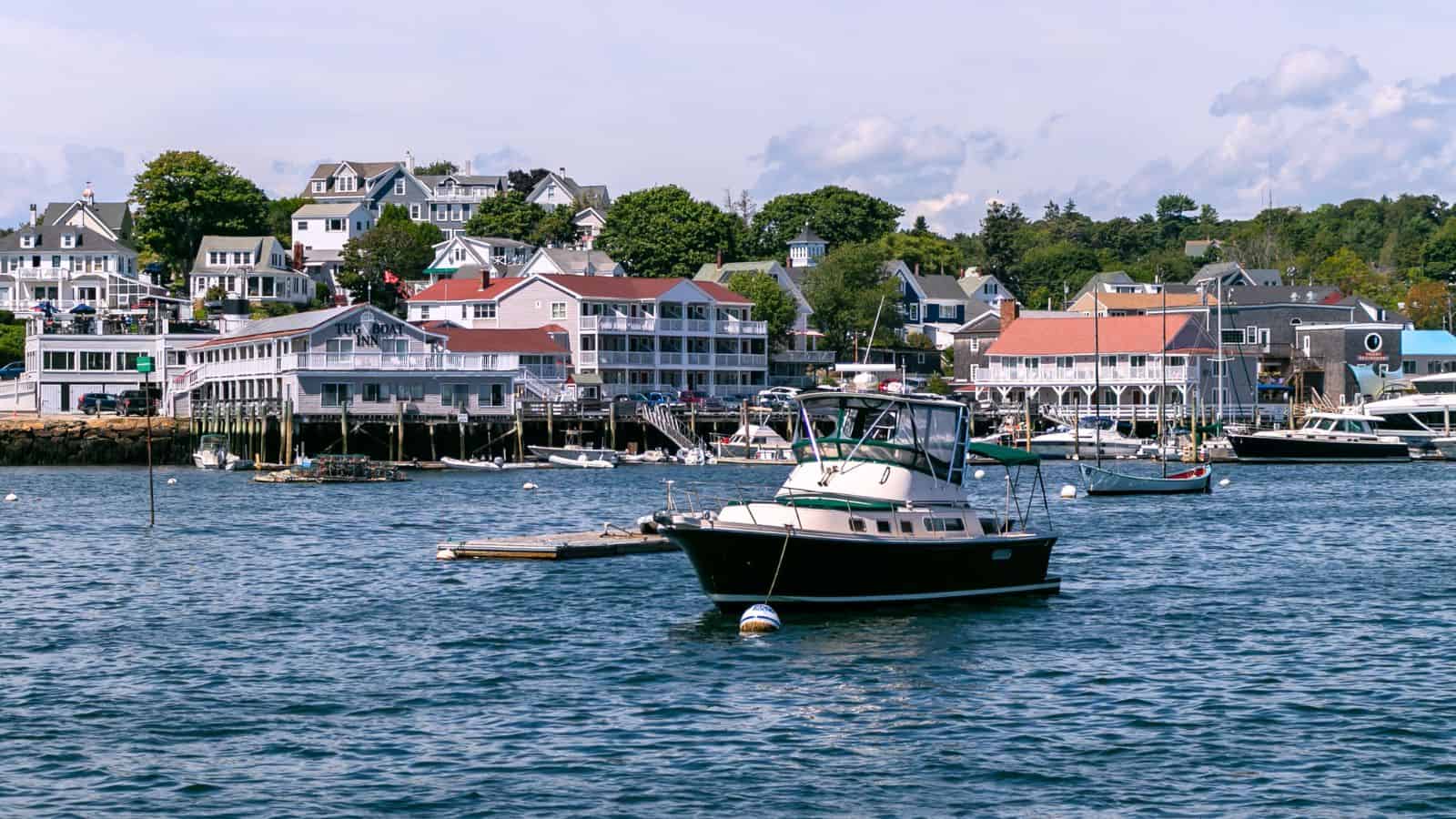 A small motorboat floats on calm water near a dock, with coastal buildings and trees lining the shore in the background under a partly cloudy sky.