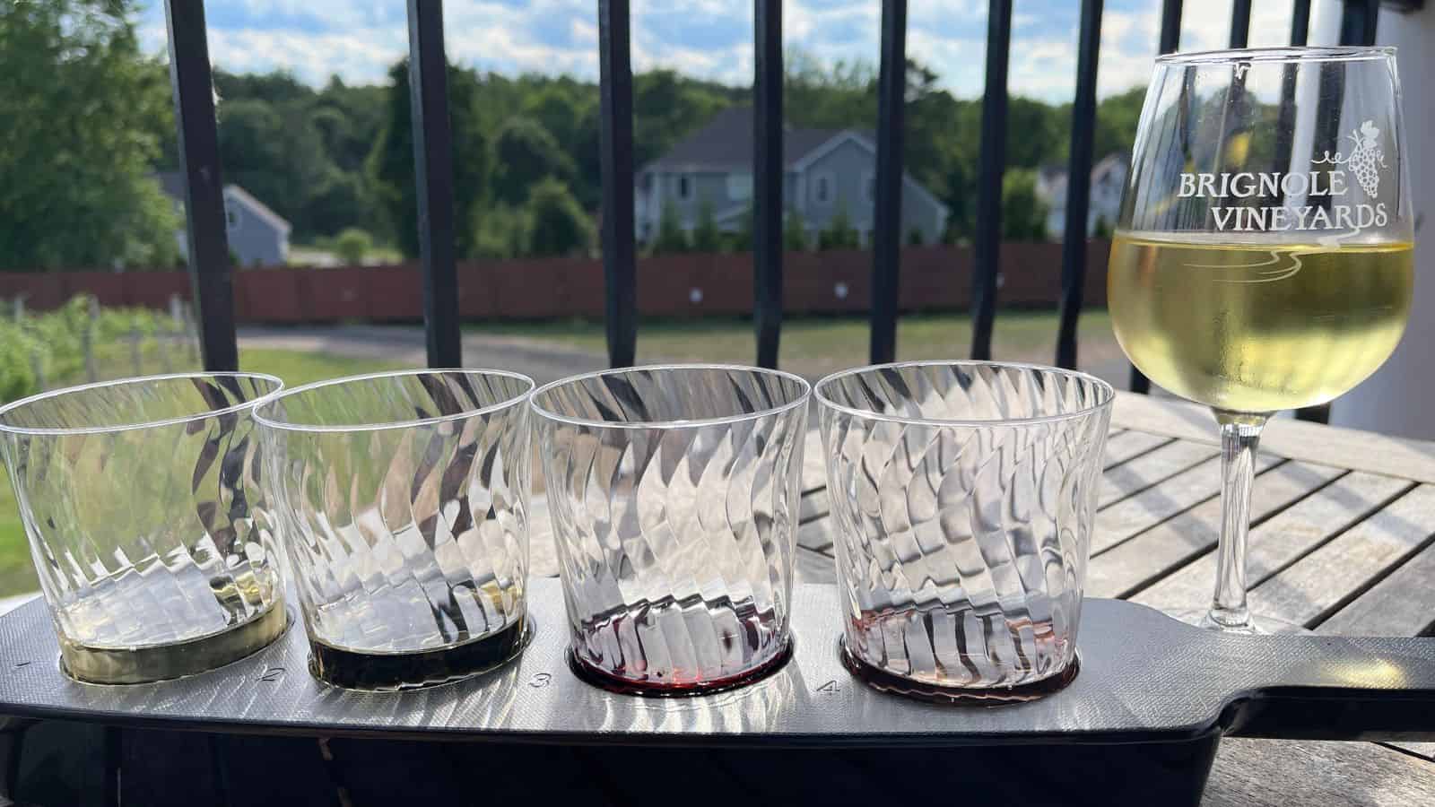 A wine flight with four empty glasses and a full glass of white wine labeled "Brignole Vineyards" on a wooden table outdoors, with houses and trees in the background.