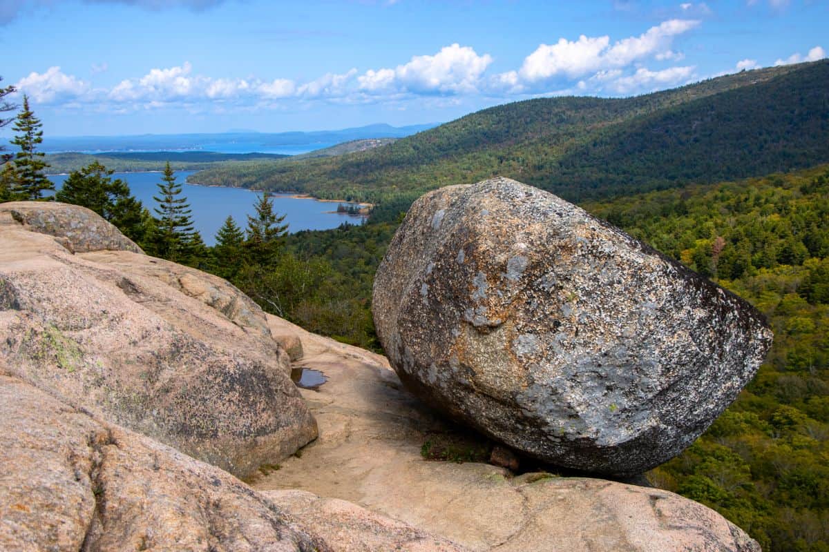 Large granite boulder resting on a rocky hilltop with forested mountains, a lake, and blue sky in the background.