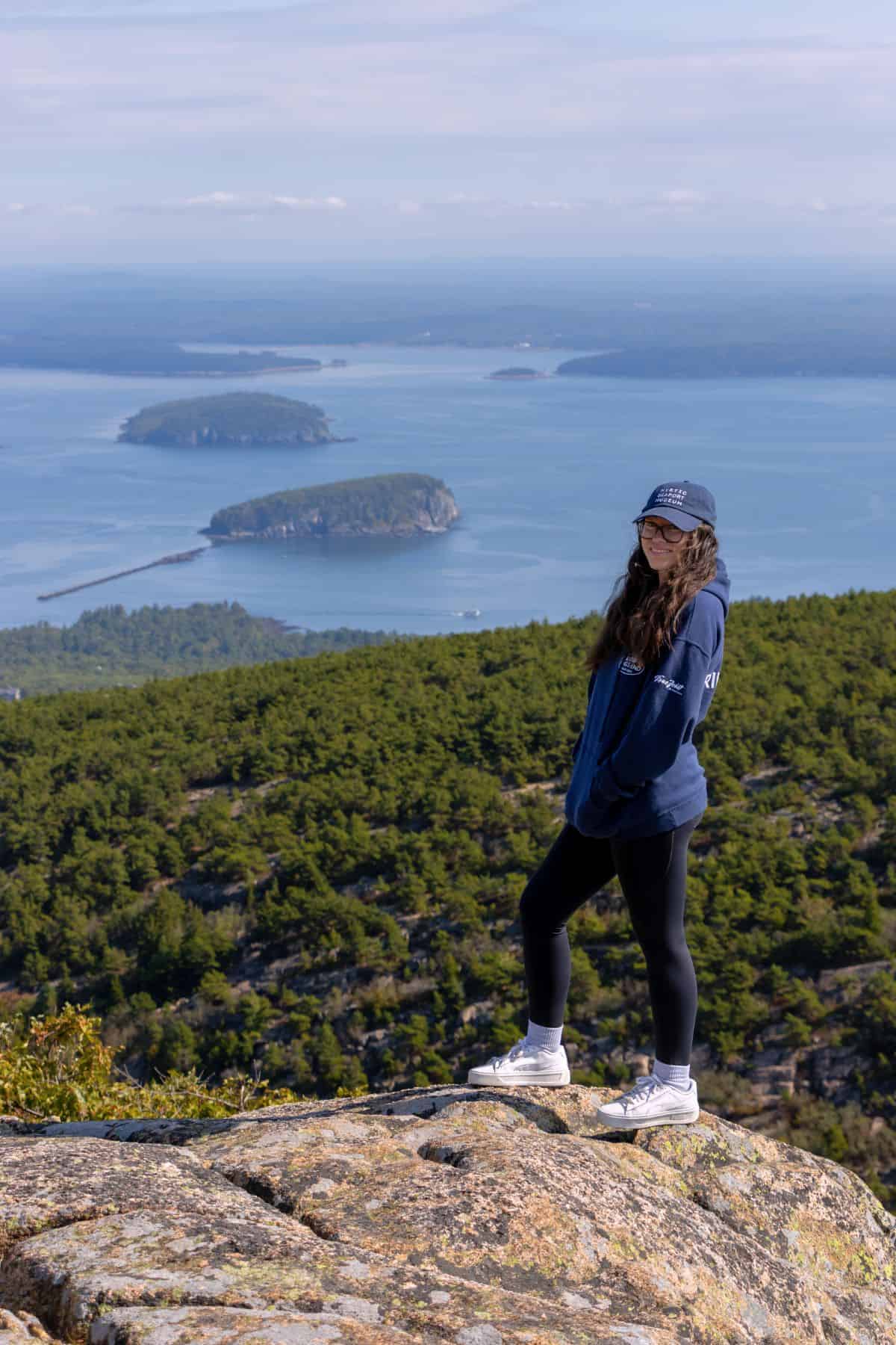 A person in a blue hoodie and black leggings stands on a rocky overlook with islands, ocean, and forested hills in the background.