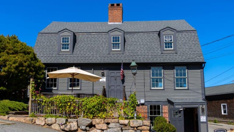 A two-story gray colonial-style house with a brick chimney, double front doors, a porch with an umbrella, and an American flag, set against a clear blue sky.