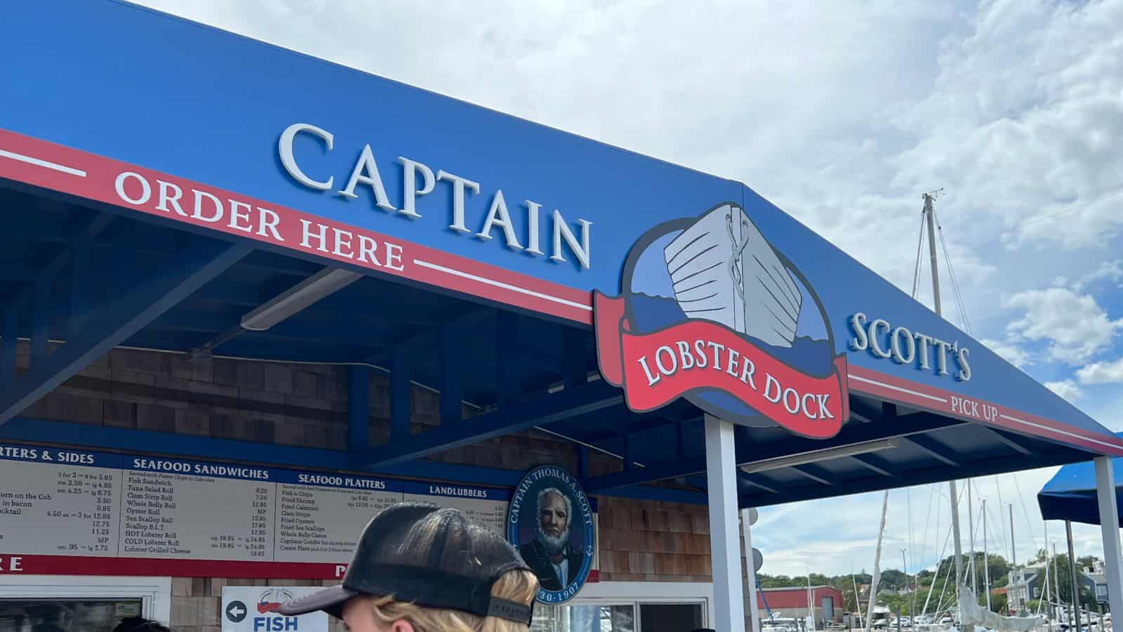 Blue and red awning at Captain Scott’s Lobster Dock, showing order and pick-up windows, menu boards, and part of a boat-filled marina in the background.