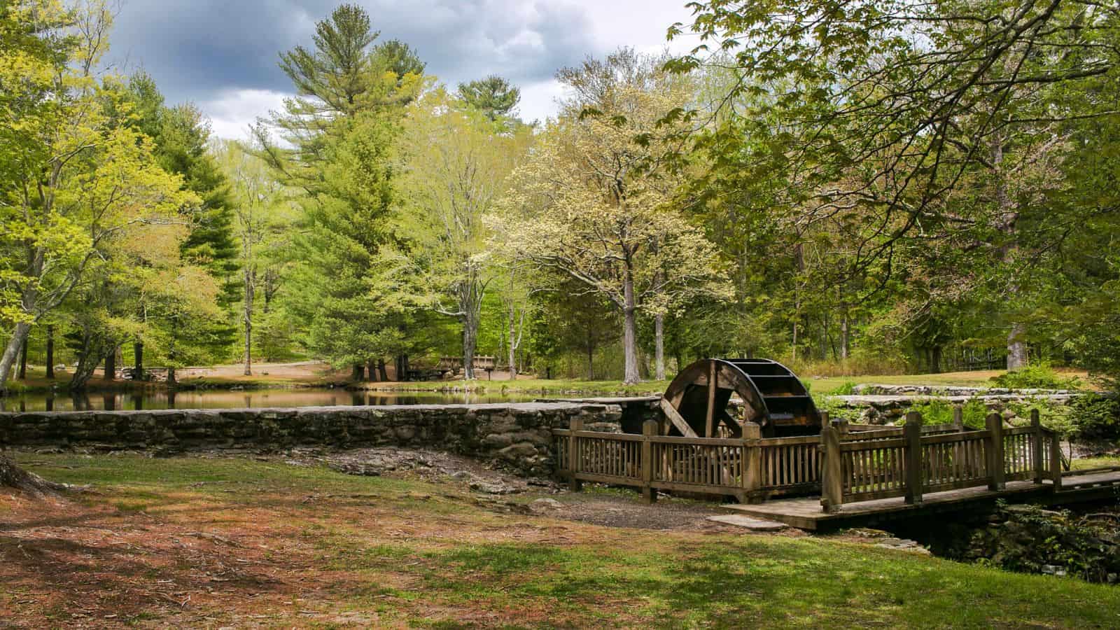 A wooden water wheel is situated by a small pond, surrounded by trees and greenery under a cloudy sky.
