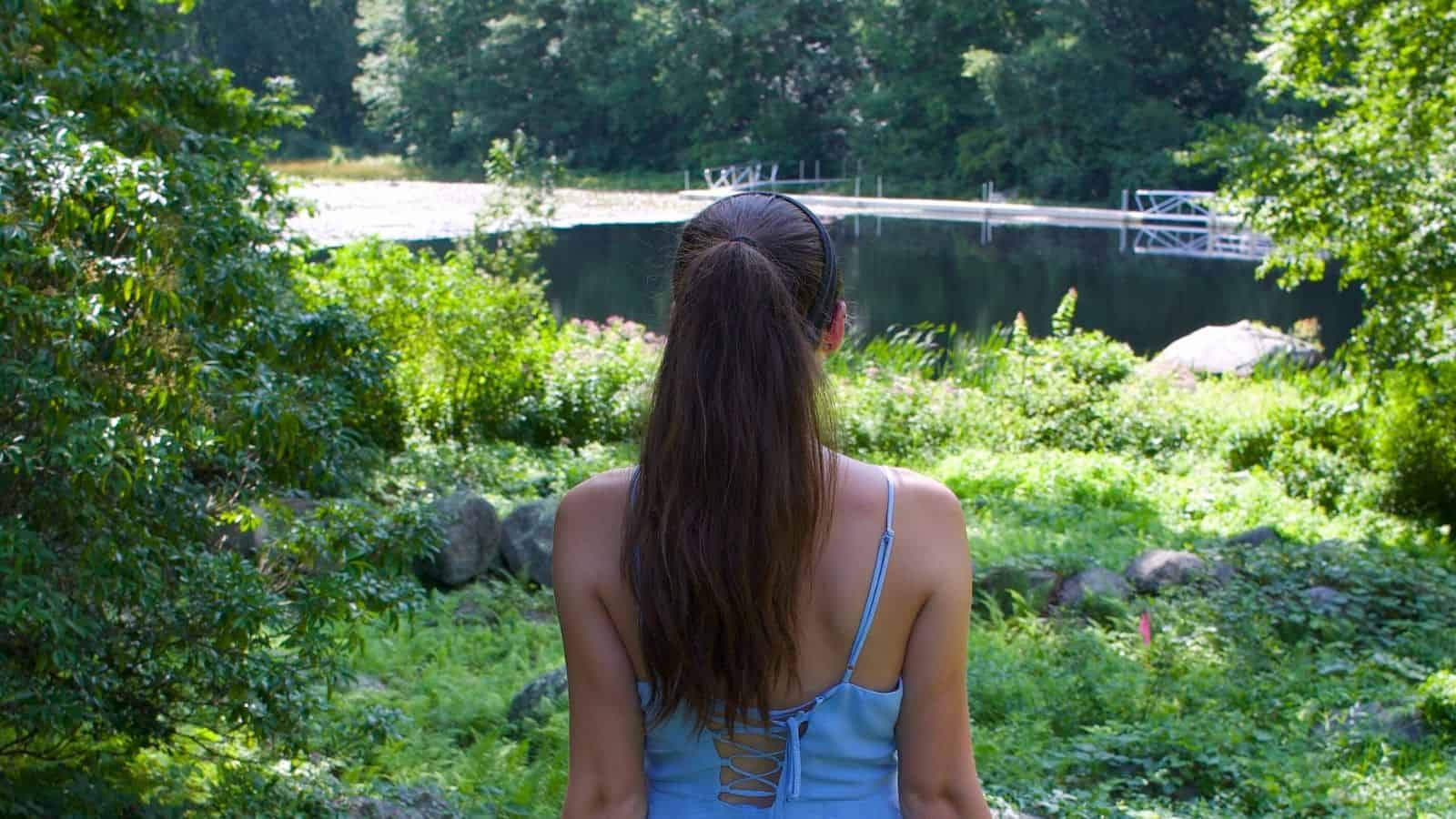 A woman with long brown hair, wearing a light blue dress, stands facing a small pond surrounded by greenery and trees.
