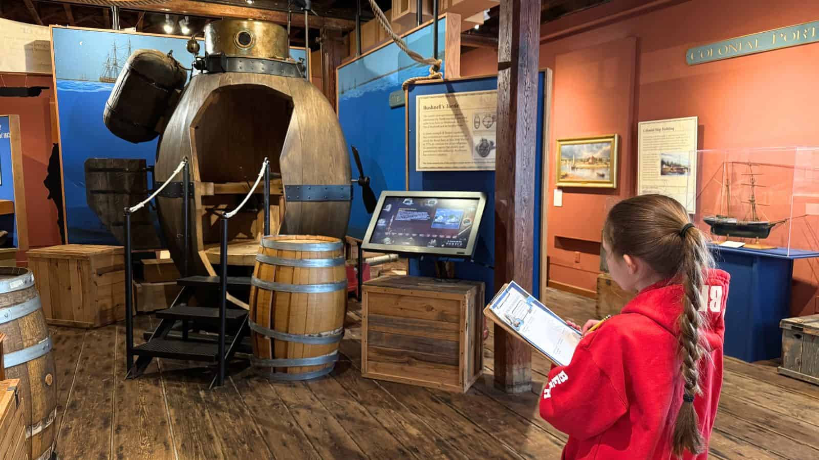A girl in a red sweatshirt holds a clipboard while observing an old submarine exhibit and barrels in a maritime museum display.