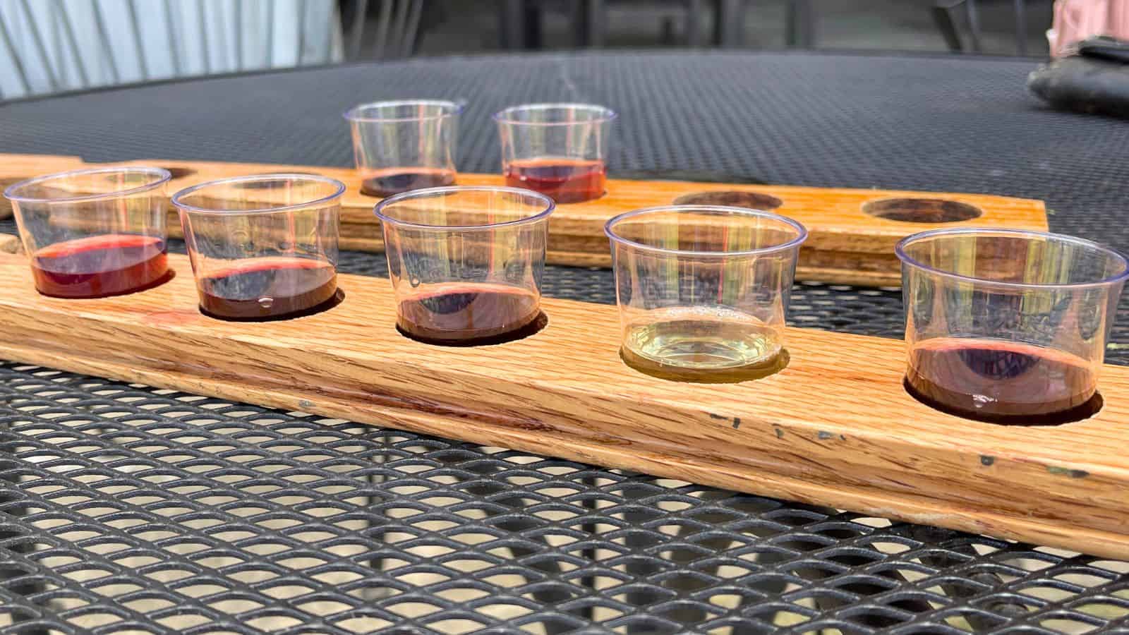 A wooden flight board holds five small plastic cups with different colored liquids, placed on a black metal mesh table outdoors.