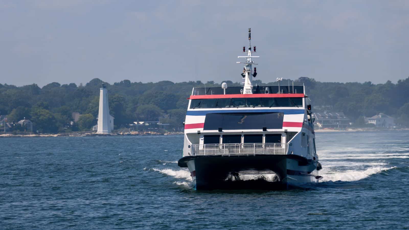 A ferry boat travels across the water toward the camera, with a shoreline, trees, houses, and a white lighthouse visible in the background.