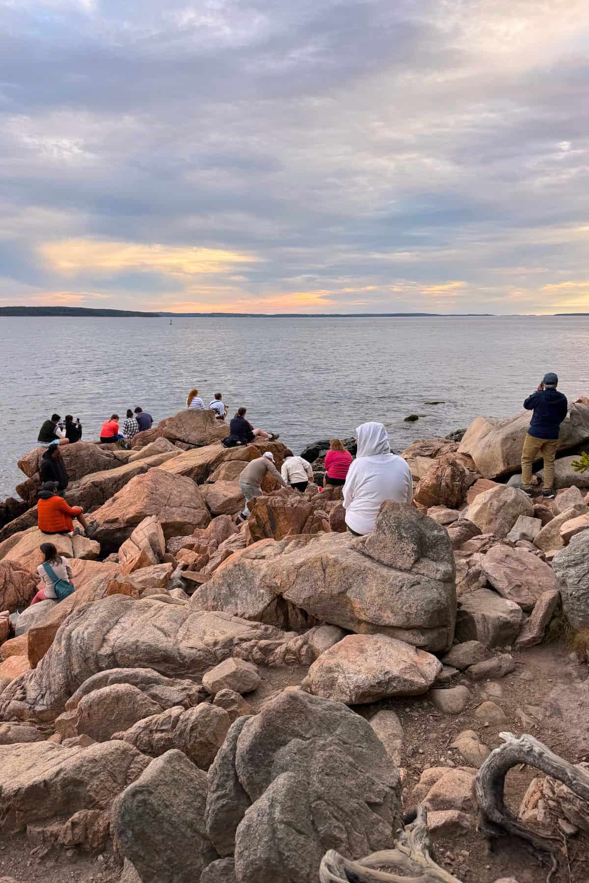 People sit and stand on large rocks by the shore, facing a calm body of water under a cloudy sky at sunset.