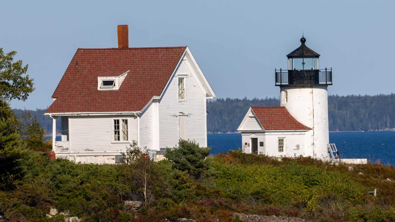 A white lighthouse with an attached small building stands beside a white house with a red roof on a green, bushy island near blue water.