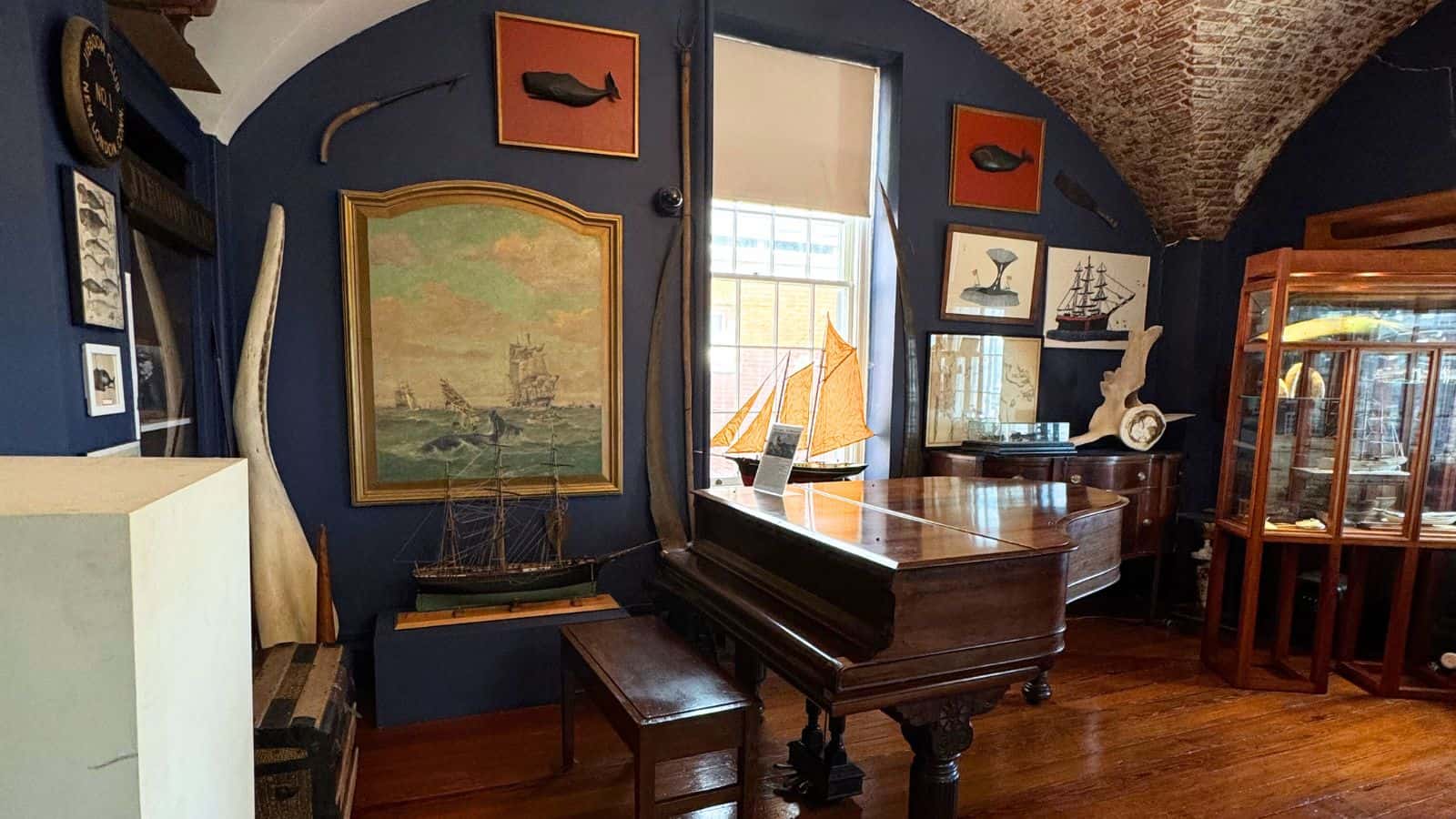 Room with a grand piano, nautical paintings, ship models, whale artifacts, and wooden floor, decorated with maritime-themed items on blue walls.