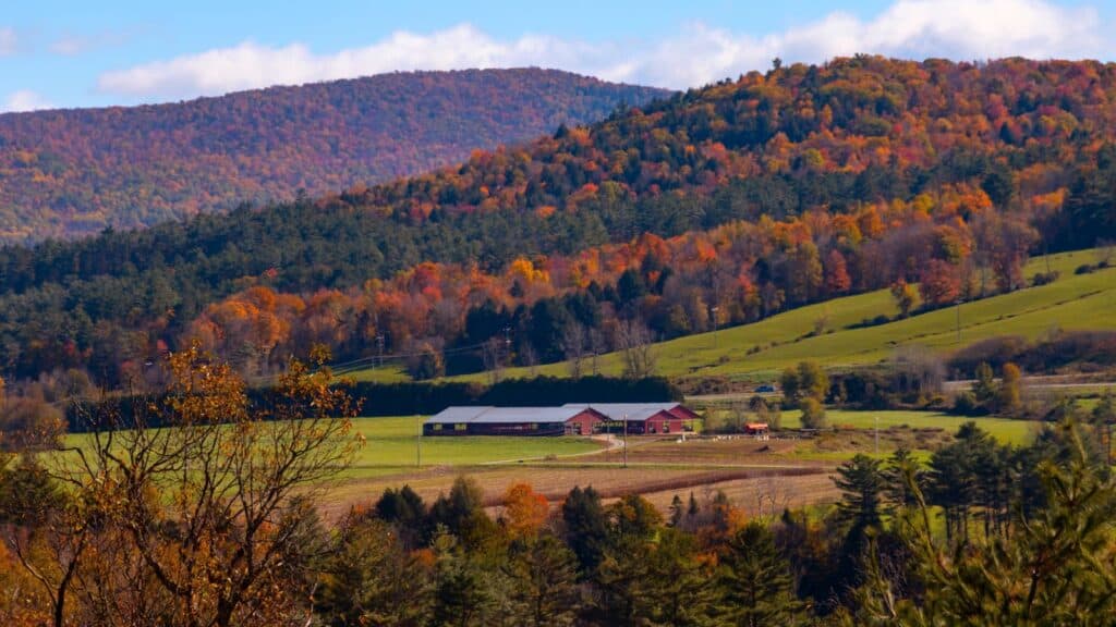 A rural landscape with a red barn and green fields surrounded by forested hills displaying autumn foliage under a partly cloudy sky.