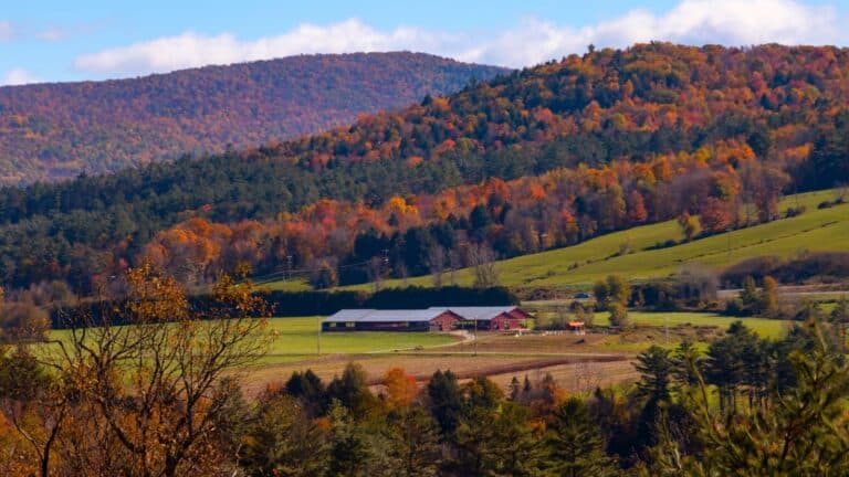 A rural landscape with a red barn and green fields surrounded by forested hills displaying autumn foliage under a partly cloudy sky.