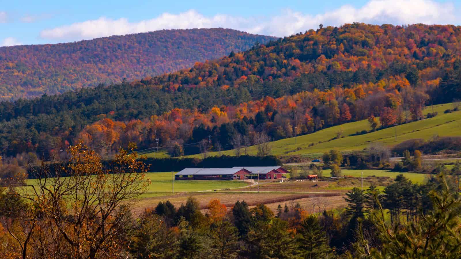 A rural landscape with a red barn and green fields surrounded by forested hills displaying autumn foliage under a partly cloudy sky.
