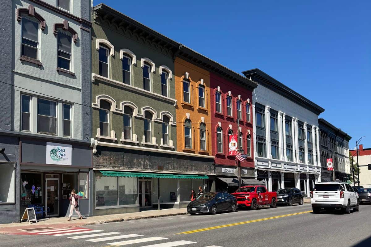 A row of colorful, historic buildings with shops and parked cars along a sunny street. Pedestrians walk on the sidewalk under a clear blue sky.