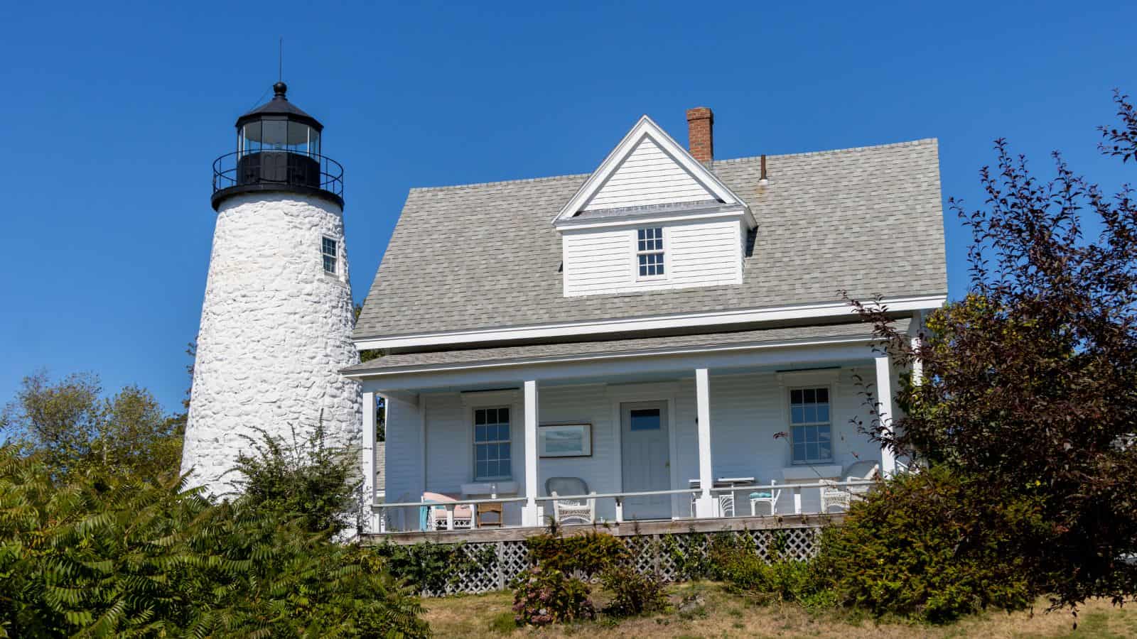 A white lighthouse with an attached house sits on a grassy hill under a clear blue sky, surrounded by bushes and trees—one of the charming lighthouses to visit in Maine.
