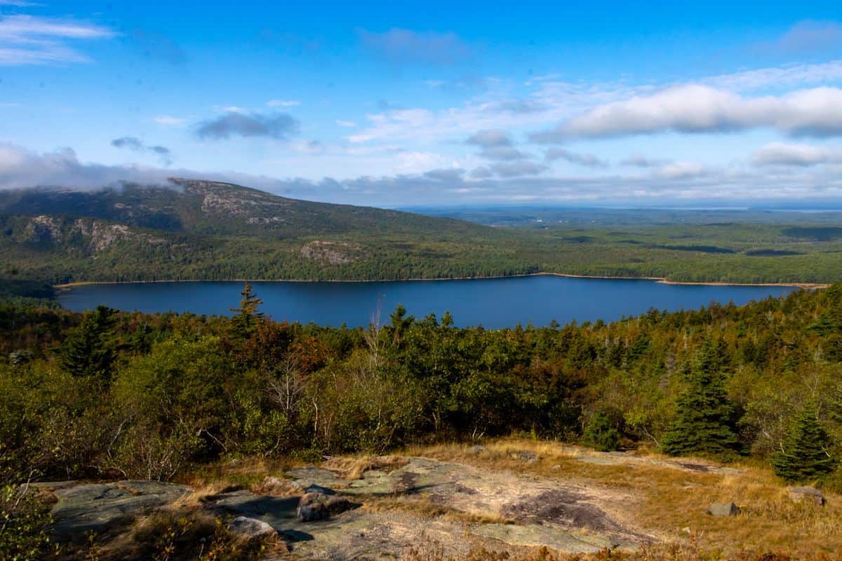 A scenic landscape with a calm lake surrounded by dense forest, distant hills, and a partly cloudy blue sky.