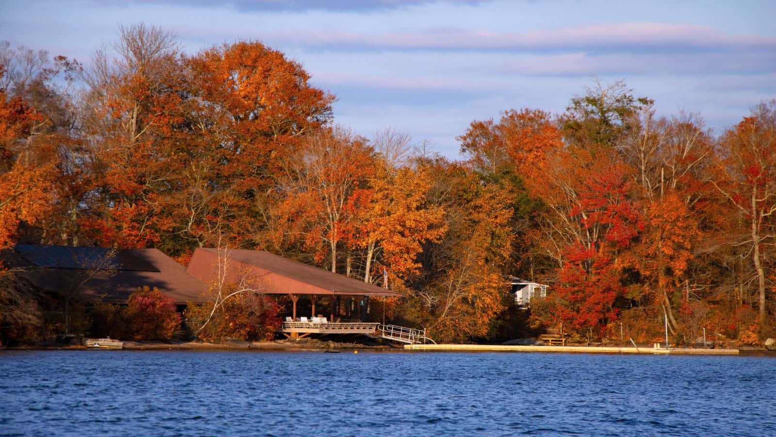 A lakeside house with a covered porch is surrounded by trees with fall foliage in shades of orange, red, and yellow under a partly cloudy sky.