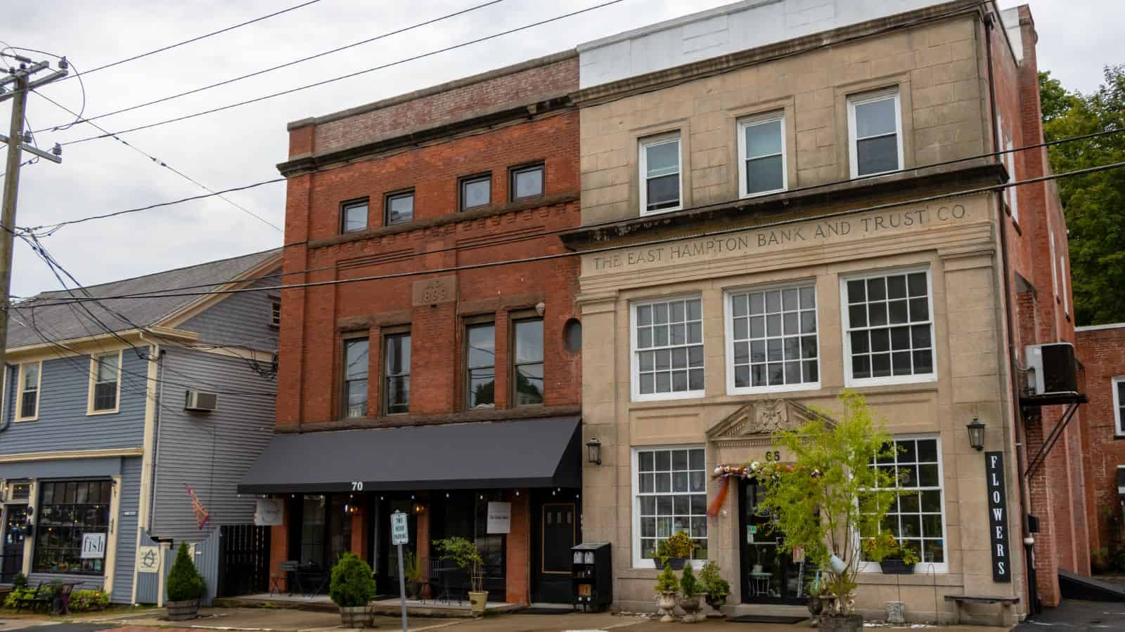 A street view of historic downtown buildings, including The East Hampton Bank and Trust Co., with storefronts and potted plants at the entrance.