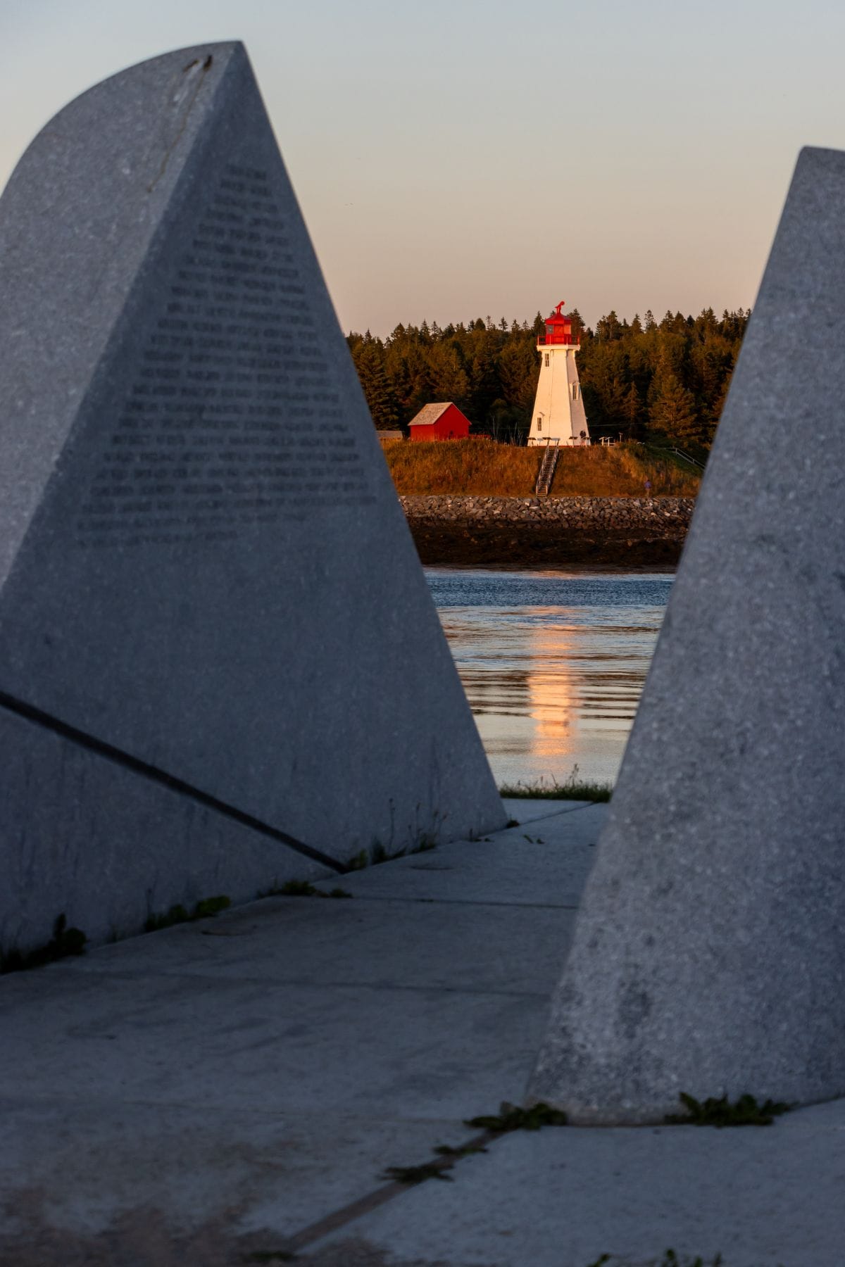 A white lighthouse with a red roof stands near a red building by the water, framed by two large stone monuments in the foreground.
