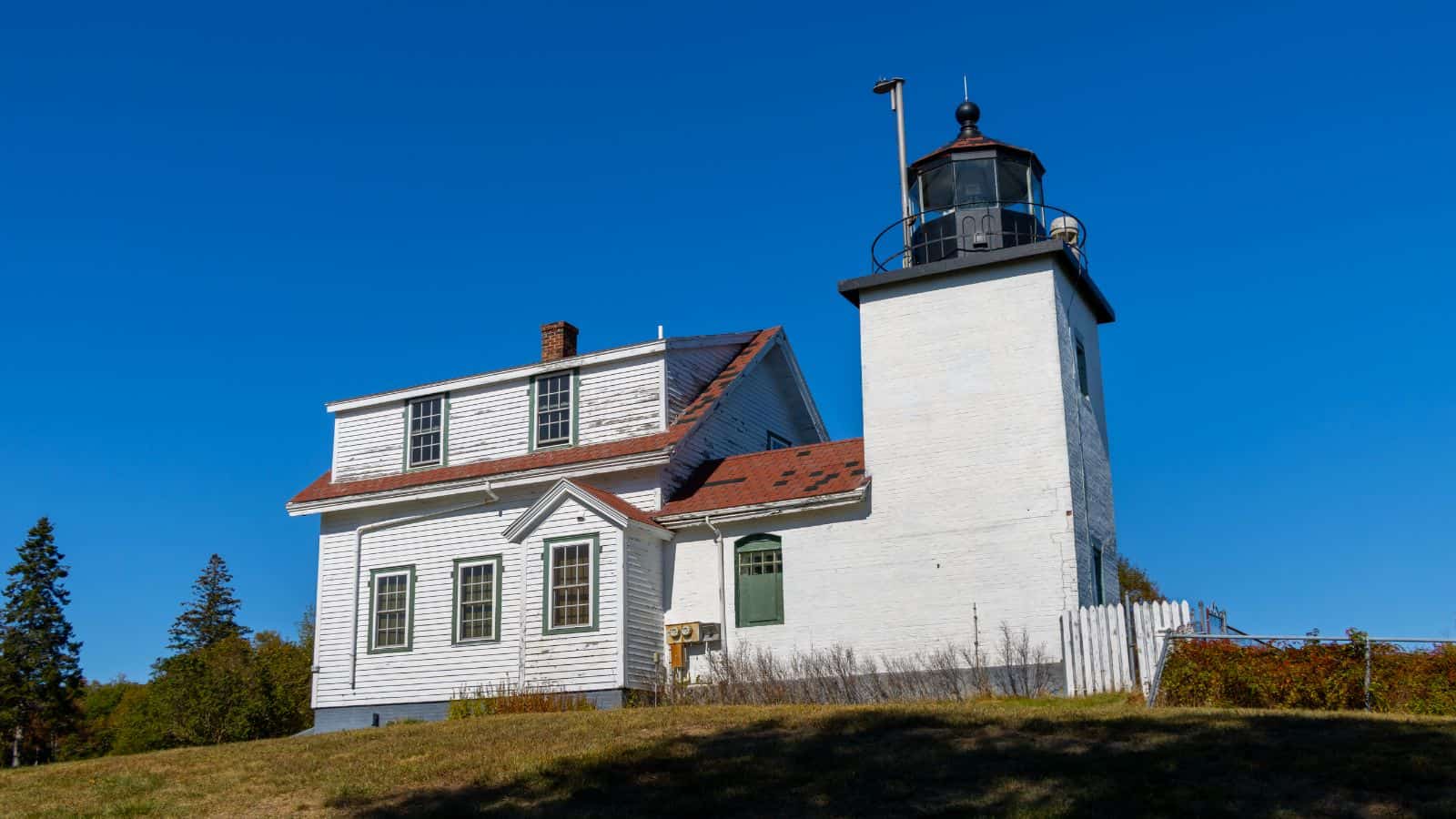 A white lighthouse with an attached keeper's house stands on a grassy hill under a clear blue sky.