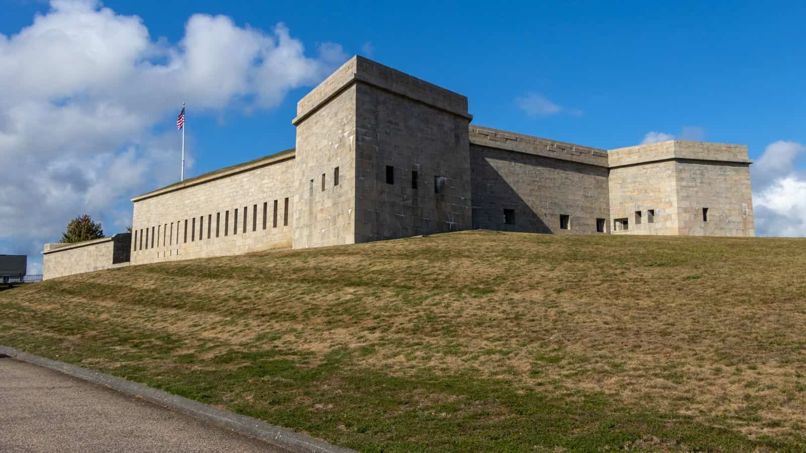 A large stone fort with rectangular towers sits atop a grassy hill under a partly cloudy sky. An American flag is flying from a flagpole.