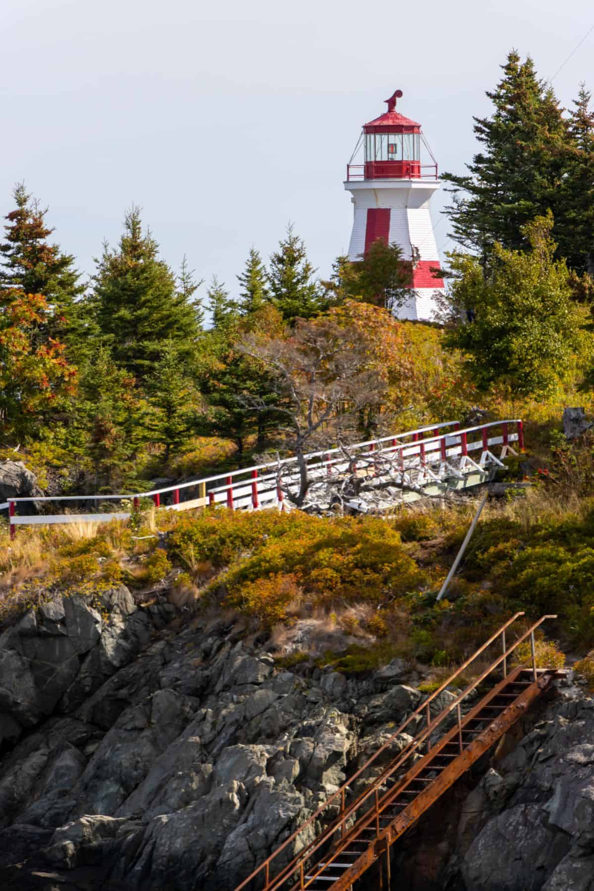A red and white lighthouse stands behind trees and a rocky hill on the border of Lubec and Campobello Island, with a wooden staircase and a white-and-red fence in the foreground.