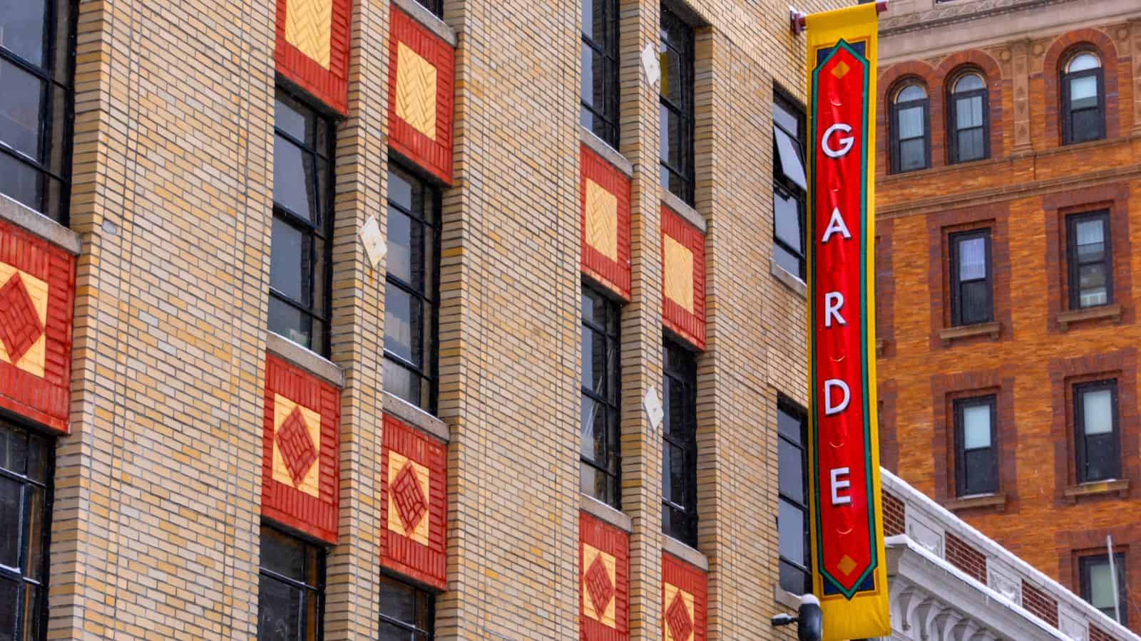 Vertical sign reading "GARDE" on the side of a tan brick building with red and yellow decorative tiles in New London, CT; another brick building is visible in the background.