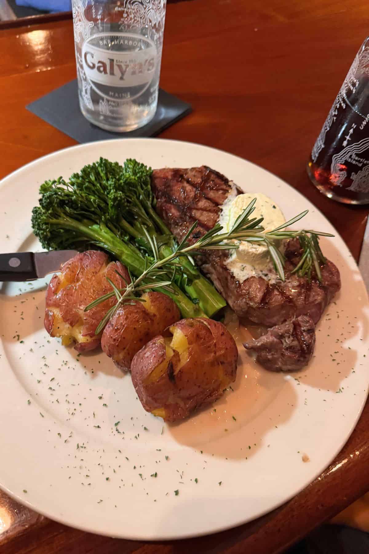 A plate with grilled steak topped with herb butter, broccolini, and three roasted smashed potatoes, garnished with fresh rosemary—perfect after a day exploring Acadia National Park in September. A knife rests on the plate; drinks are in the background.