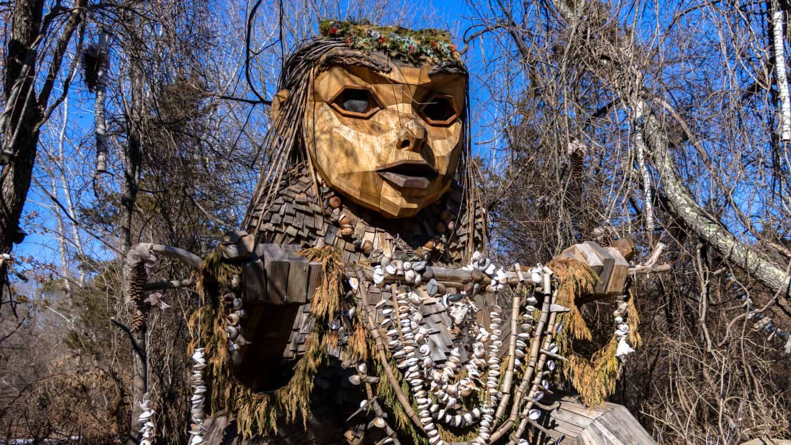 Large wooden sculpture of a figure with long hair, shell necklaces, and leafy decorations, set outdoors among leafless trees under a clear blue sky.