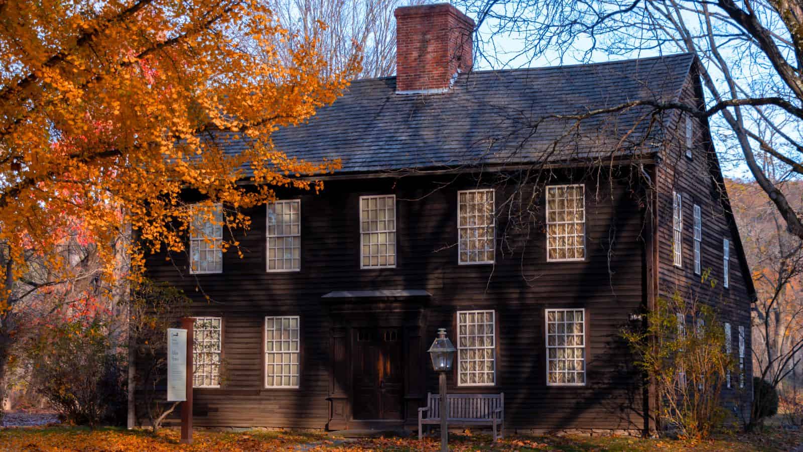 Two-story, dark wooden house with many paned windows and a central brick chimney, surrounded by autumn trees and fallen leaves.