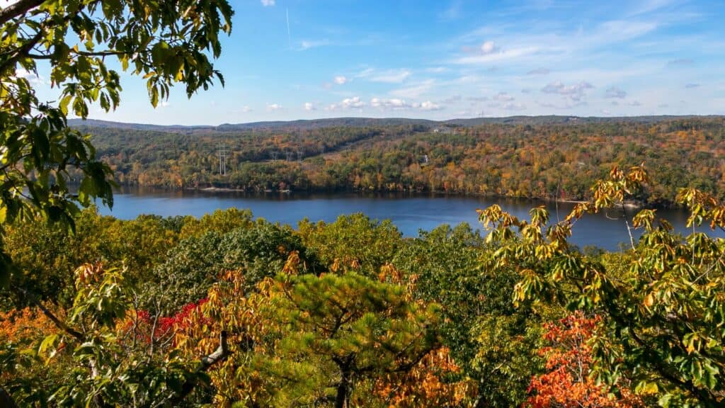 A river runs through a forested landscape with trees displaying autumn colors under a partly cloudy sky.