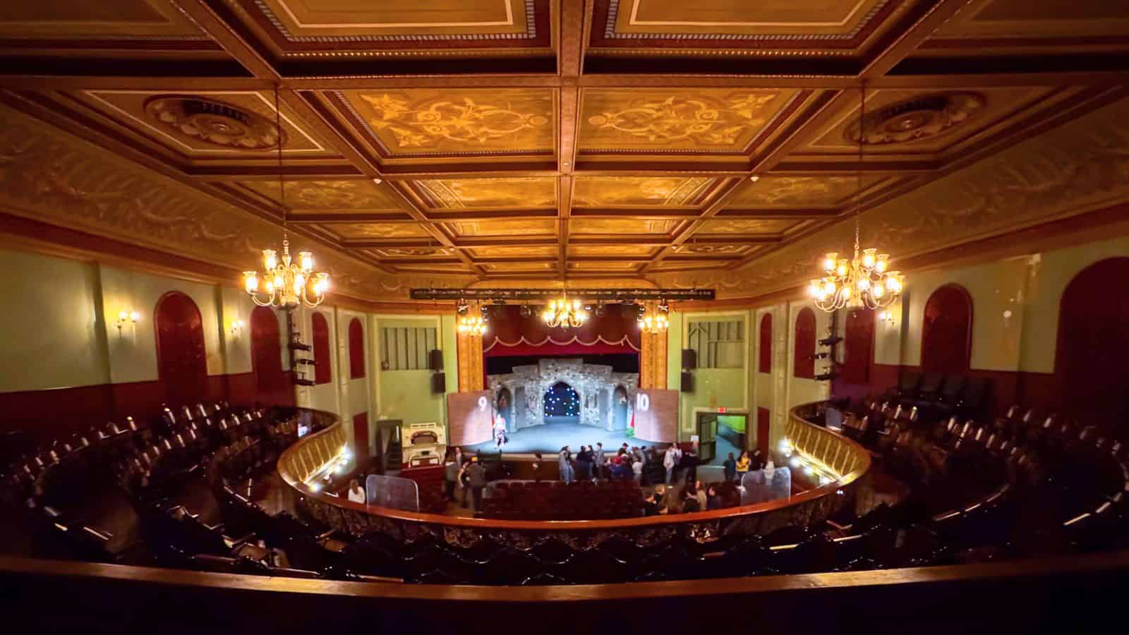 Ornate theater interior with chandeliers, decorative ceiling, and a stage set for a performance; people gather near the stage of one of the most intriguing haunted places in Connecticut, while seats remain mostly empty.