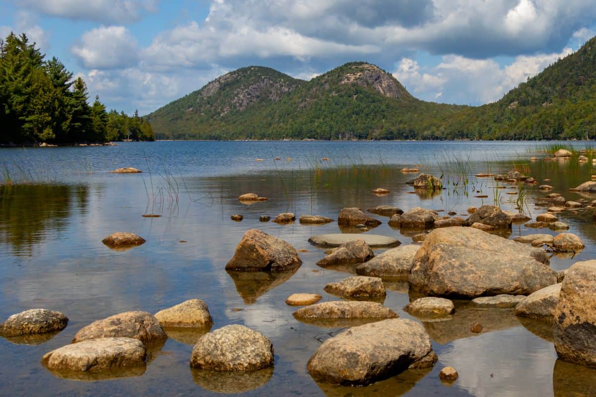 A calm lake with scattered rocks in the foreground, surrounded by forested hills under a partly cloudy sky.