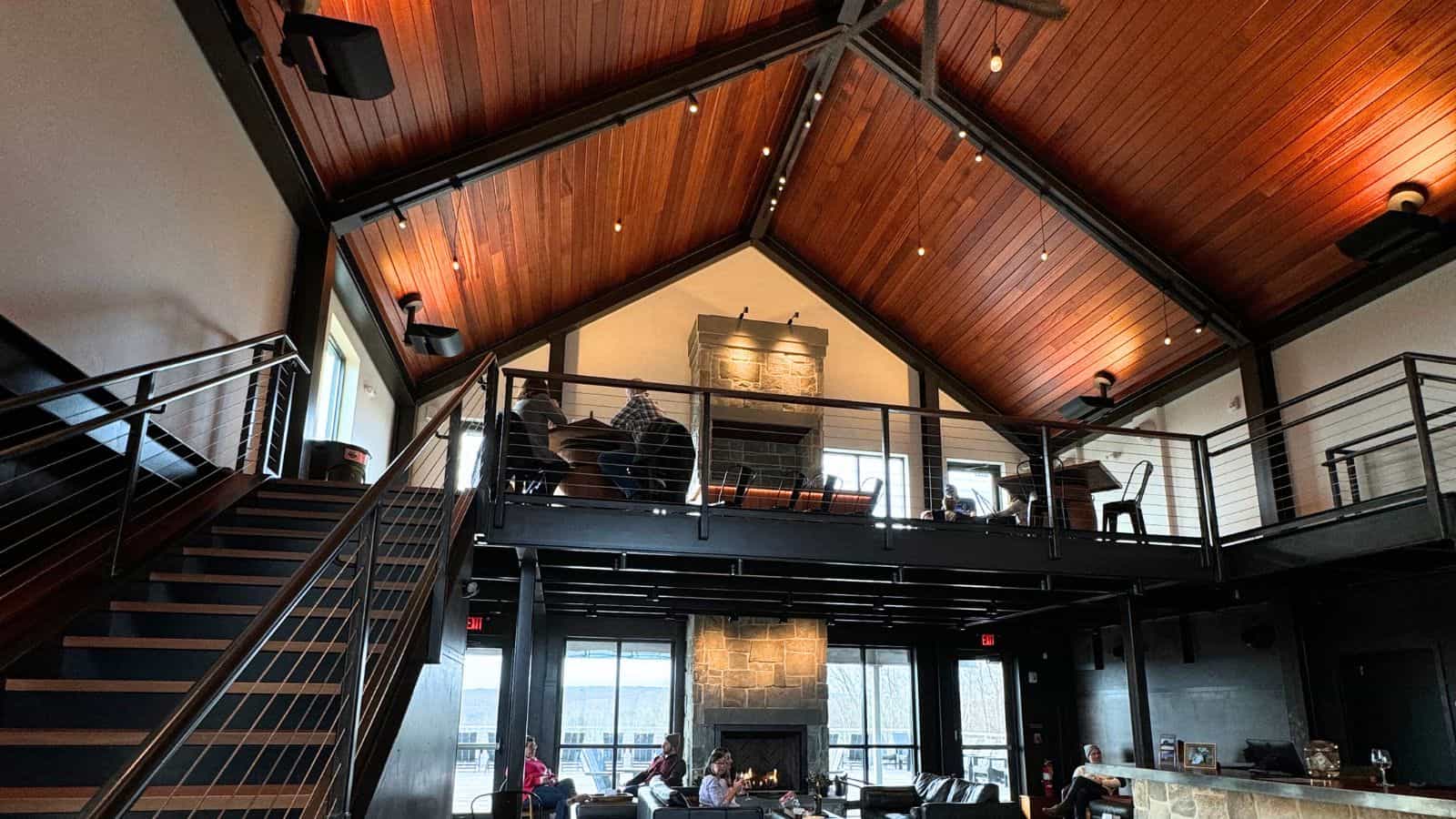 Spacious modern lounge with wood ceiling, metal railings, large windows, and a stone fireplace. People are seated on both the ground floor and upper mezzanine.