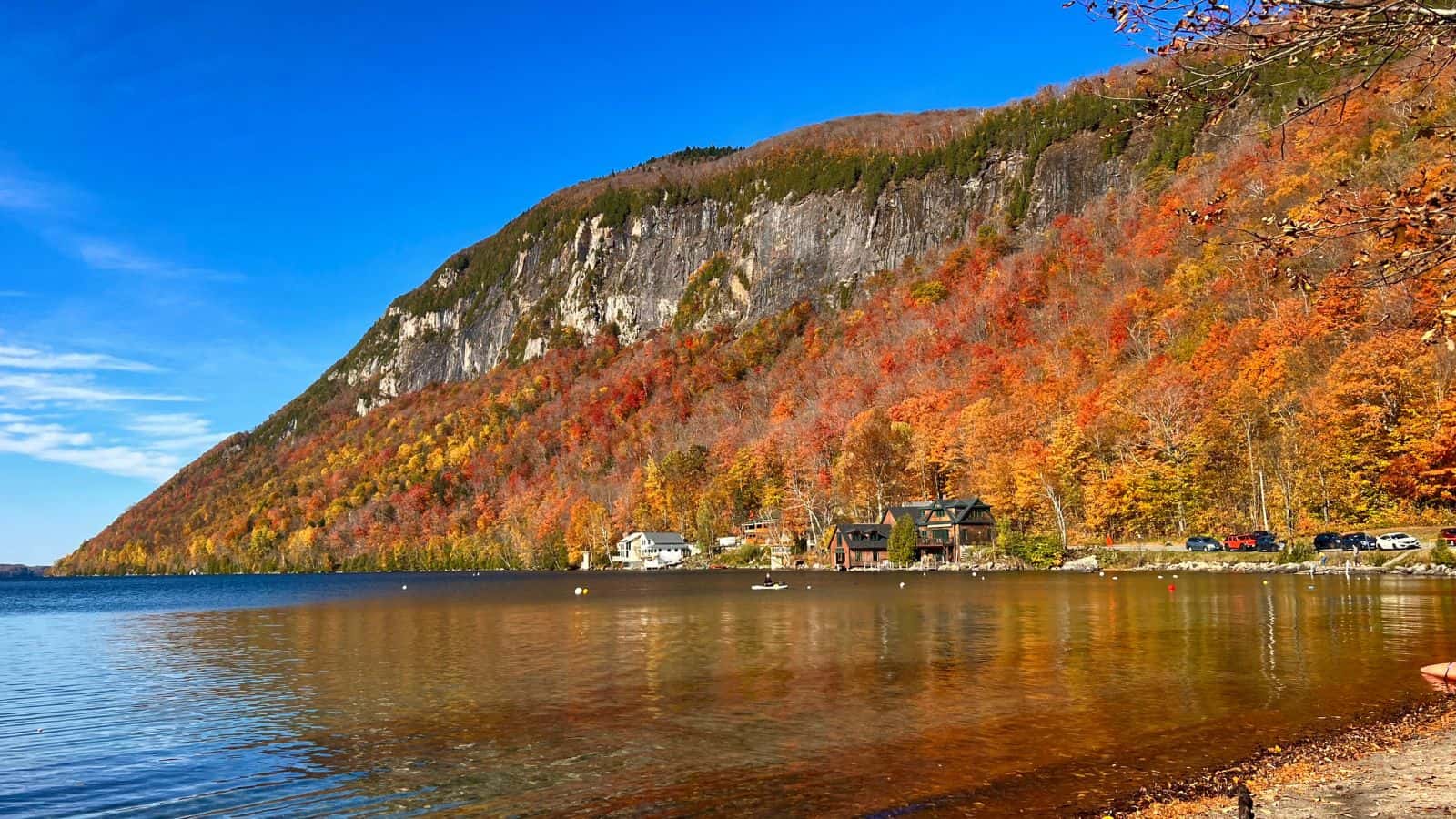 A calm lake bordered by autumn-colored trees and a steep, rocky hill, with a few houses and blue sky in the background.