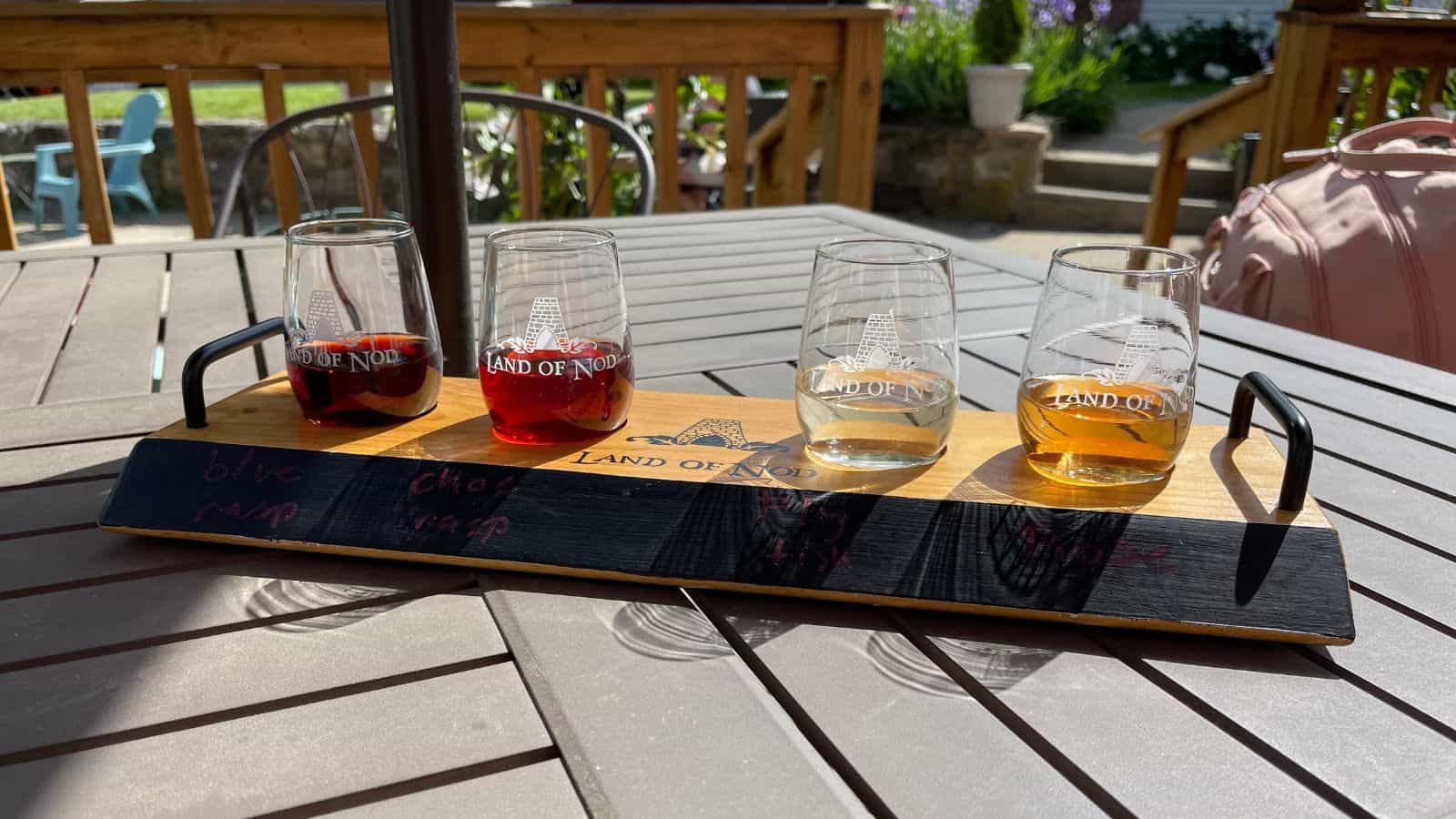 Four glasses of wine arranged on a wooden flight board with labels, sitting on an outdoor patio table in sunlight.
