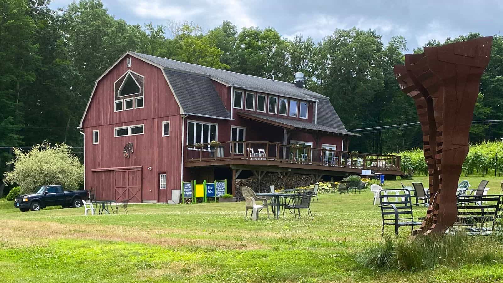 A red barn-style building with a deck overlooks a grassy area with patio chairs, a metal sculpture, a parked truck, and trees in the background.