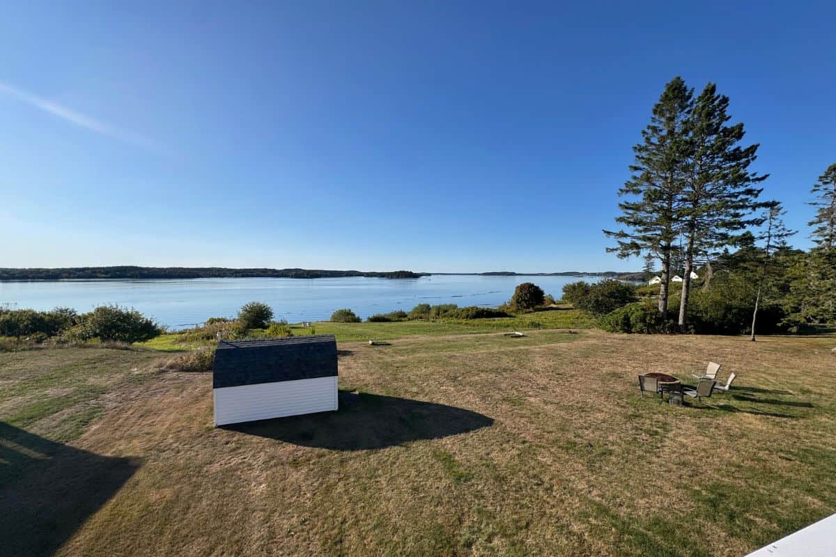A small shed sits on a grassy lawn near a waterfront, with outdoor chairs and a fire pit nearby; clear blue sky and trees in the background.