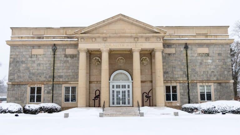 A neoclassical stone building with four columns at the entrance, labeled "Lyman Allyn Museum," stands in new london ct, surrounded by snow-covered ground and bushes.