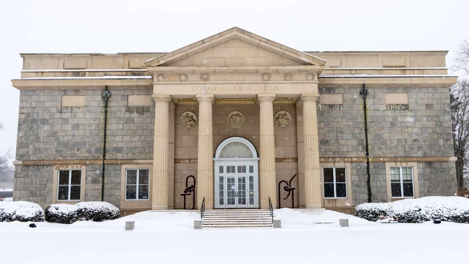 A neoclassical stone building with four columns at the entrance, labeled "Lyman Allyn Museum," stands in new london ct, surrounded by snow-covered ground and bushes.