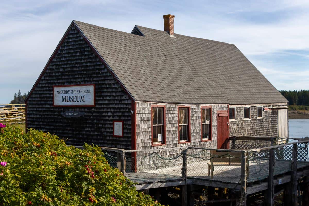 A wooden shingle-sided building with a sign reading "McCurdy Smokehouse Museum" stands by the water, surrounded by greenery.