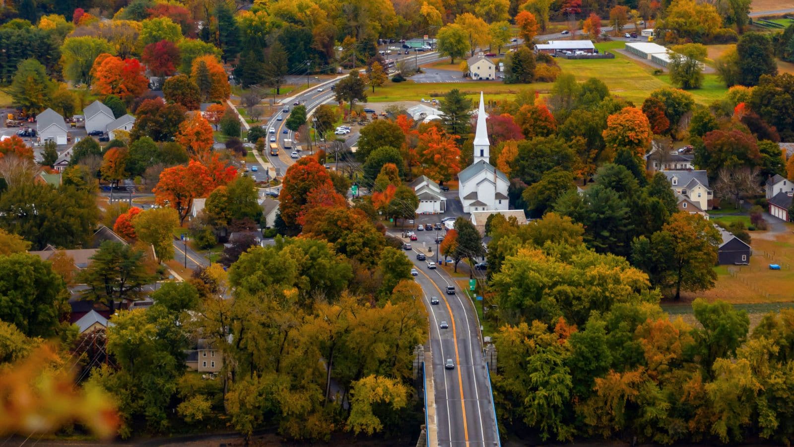 A small town with a white church, houses, and roads surrounded by trees with autumn foliage in shades of red, orange, and yellow.