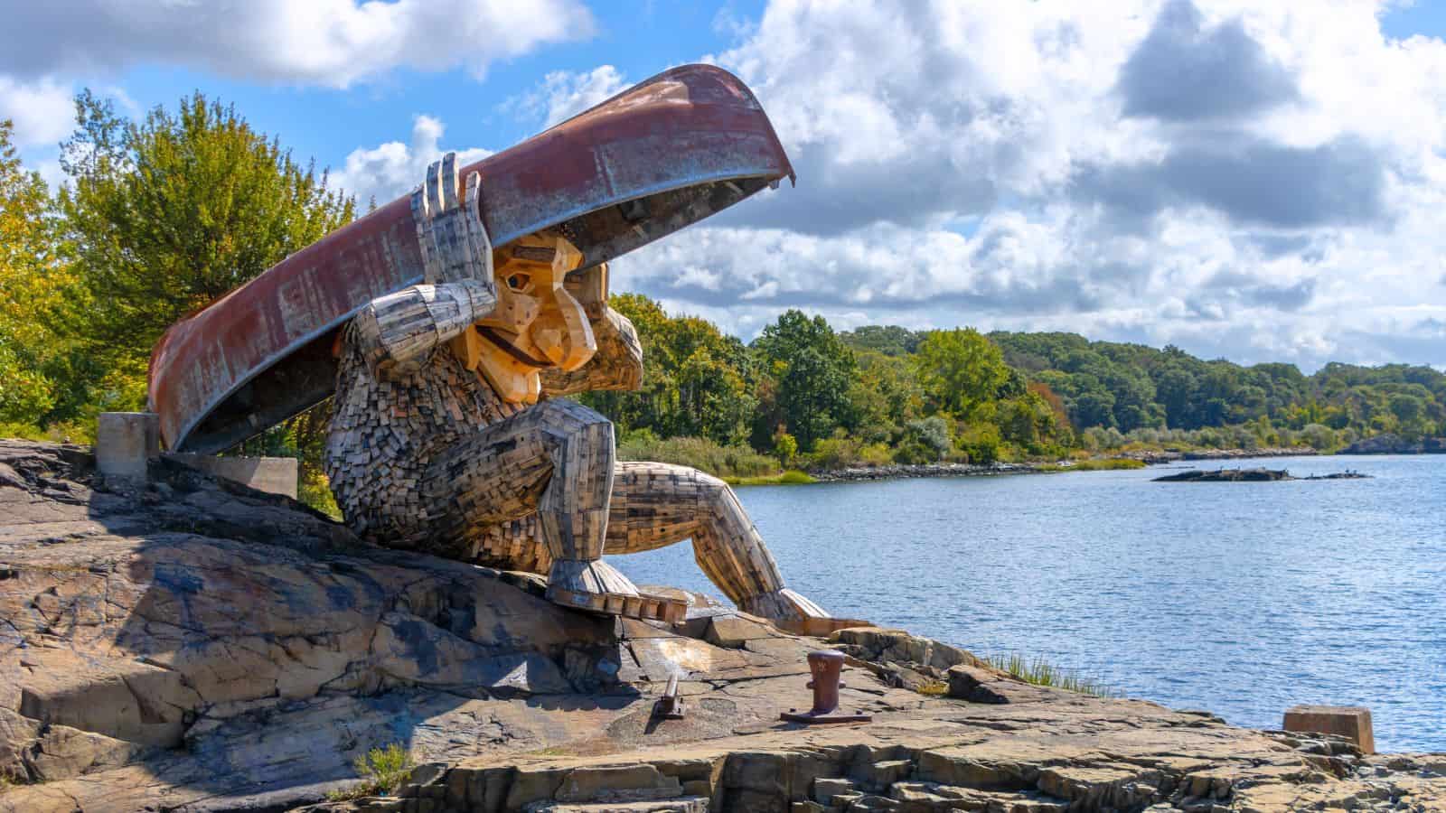 A large wooden troll sculpture sits on rocks by the water, holding a canoe over its head, with trees and water in the background.